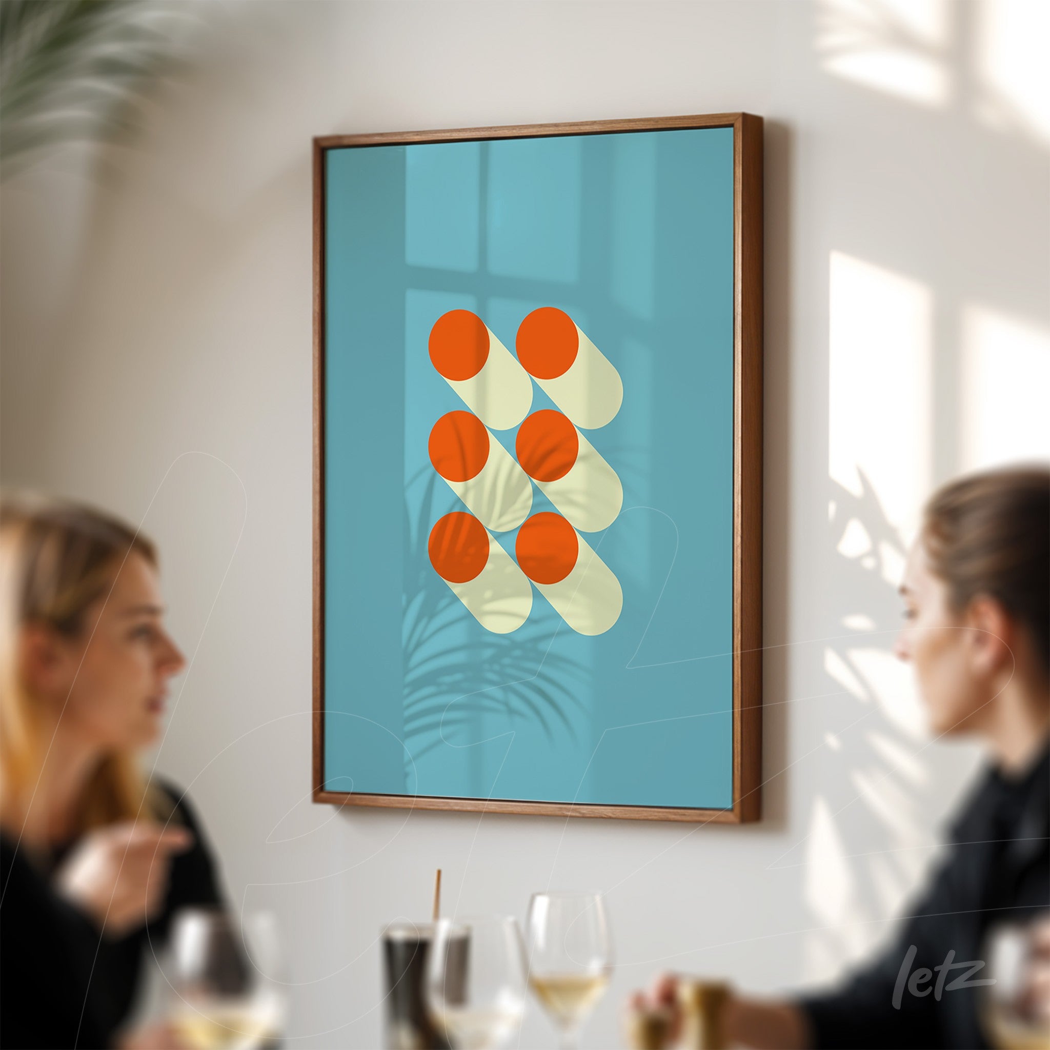 quadro com arte geométrica de círculos laranja e sombra amarelo pálido sobre fundo azul, emoldurado em madeira escura, exibido em parede branca com duas pessoas ao redor