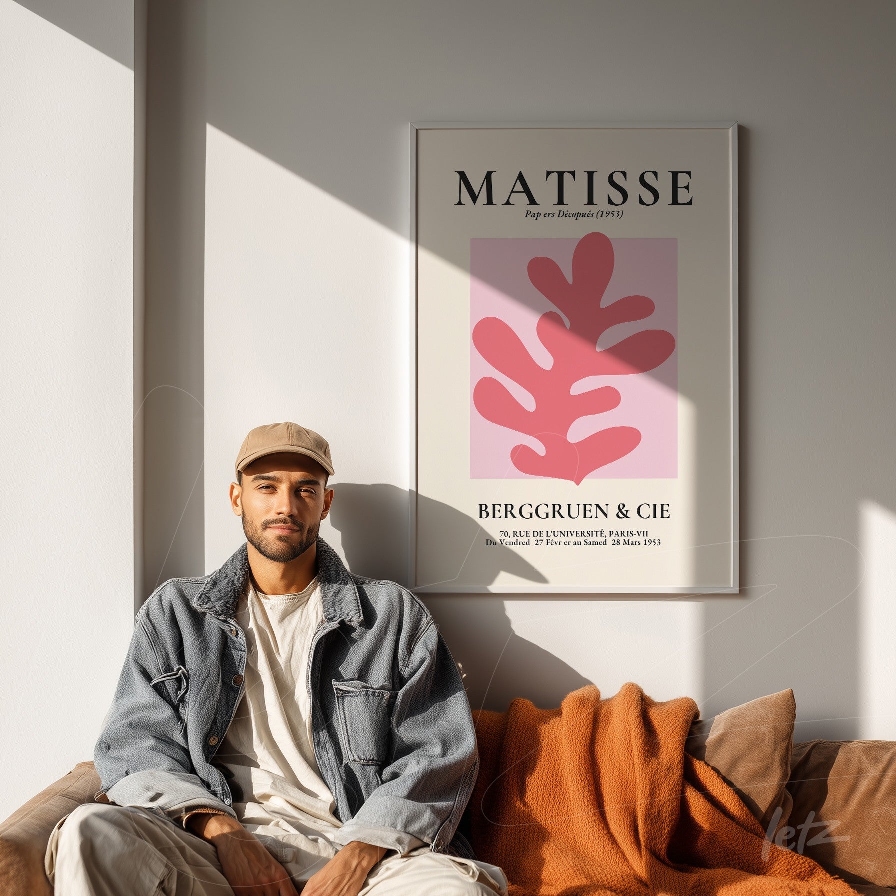 quadro com arte moderna de fundo rosa e folhas em tons de vermelho, moldura branca, exposto em parede iluminada com sofá marrom e manta laranja