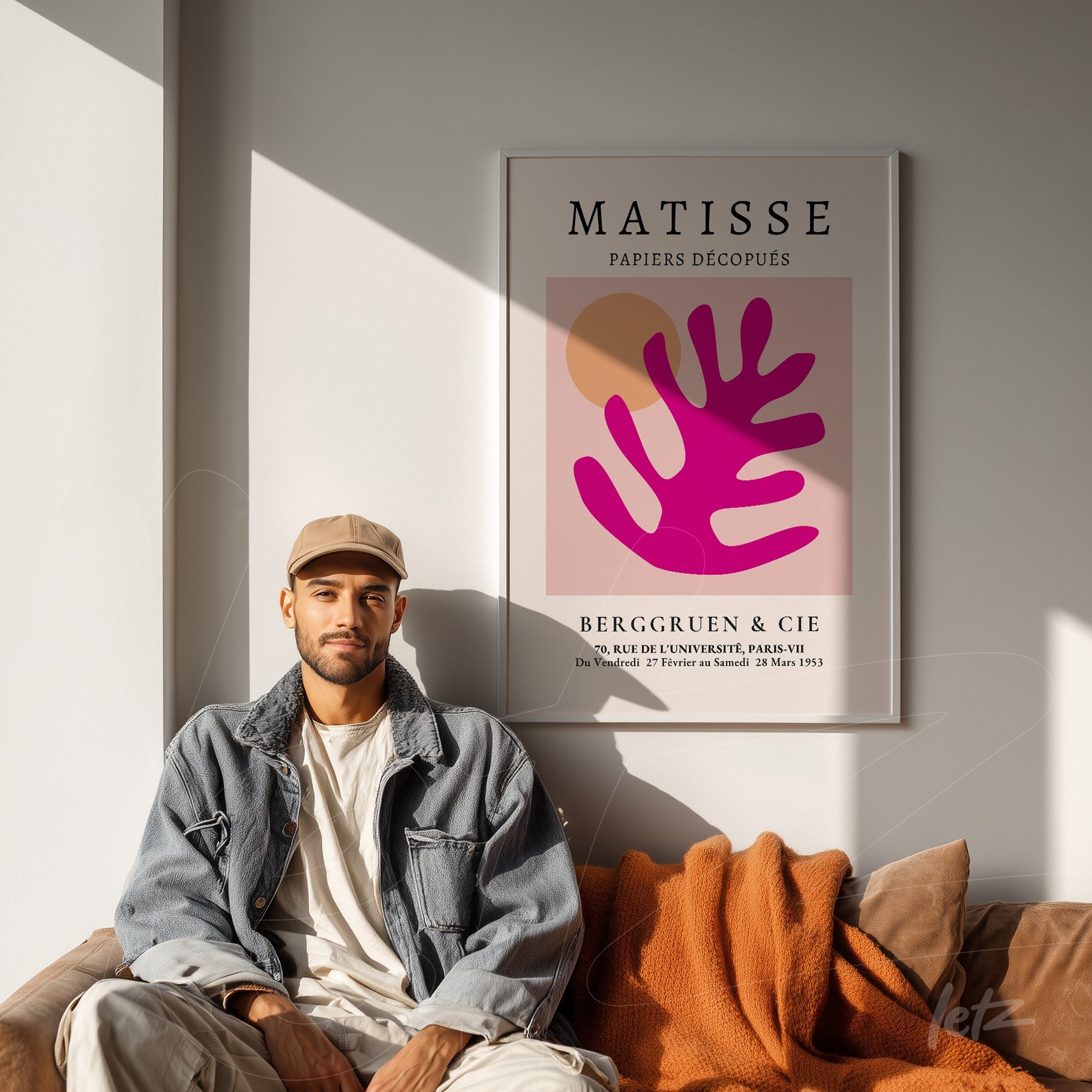 quadro emoldurado com arte de Matisse em tons de rosa e laranja, exibido sobre uma parede clara, com um homem sentado em um sofá cobrindo-se com um lençol laranja