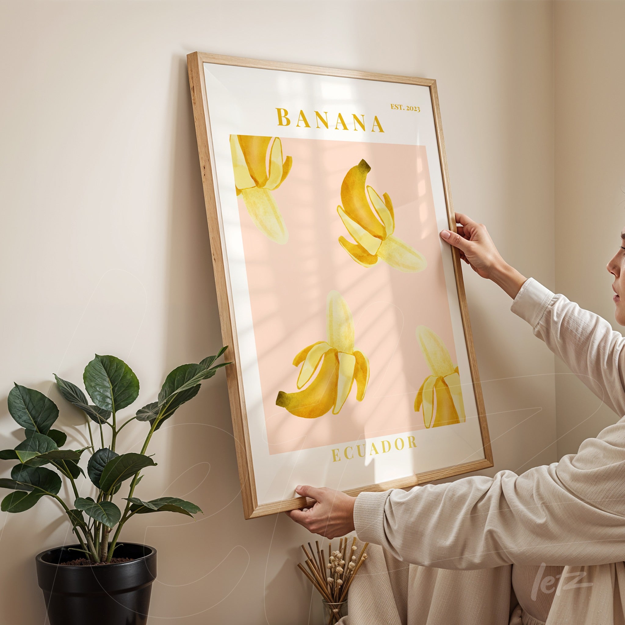 quadro emoldurado com ilustração de bananas amarelas sobre fundo rosa, segurado por uma mulher frente a uma planta