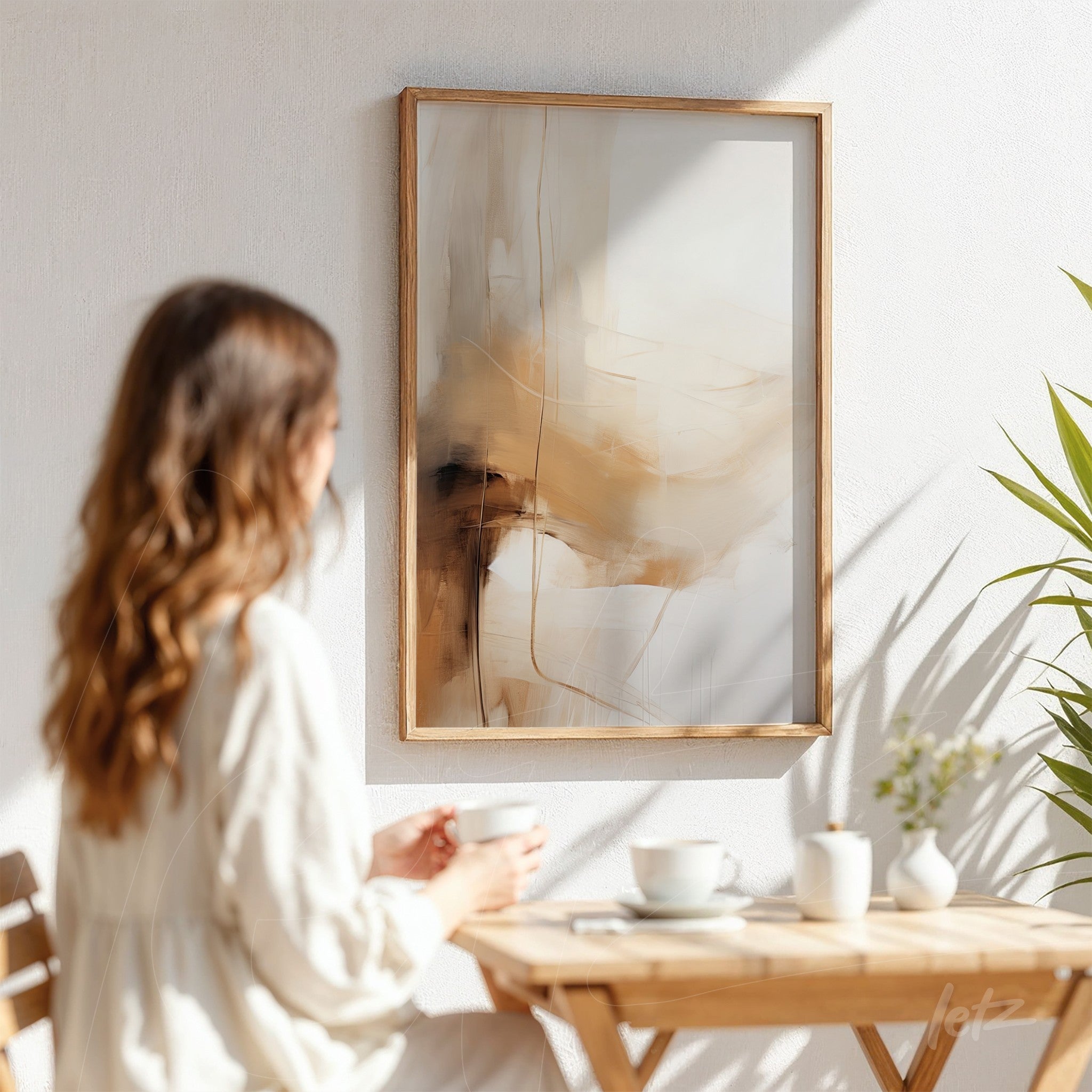 quadro abstrato em moldura de madeira clara, com tons neutros de bege e marrom, exposto em parede branca enquanto uma mulher senta em cadeira de madeira