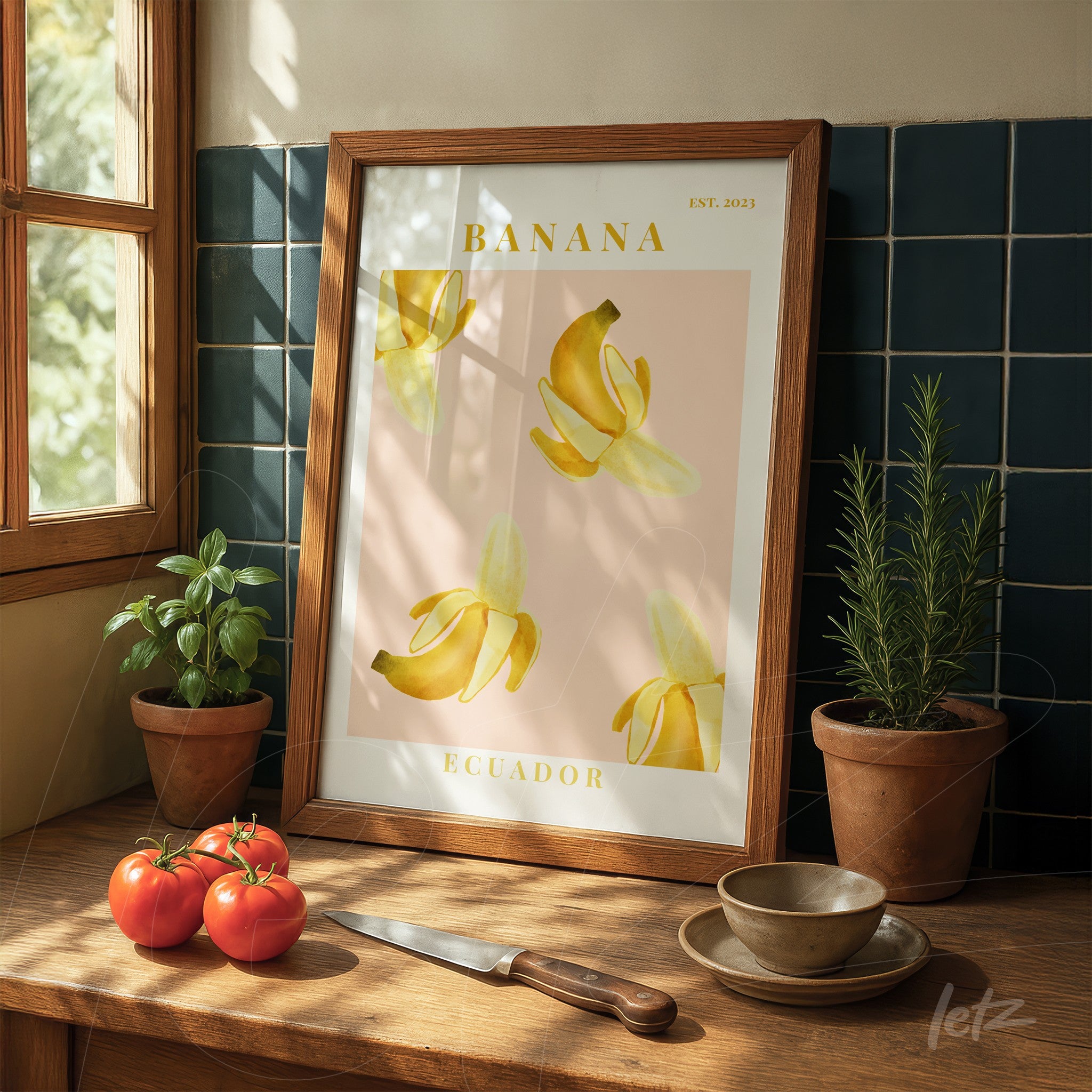 quadro emoldurado com ilustração de bananas sobre fundo rosa, apoiado em mesa de madeira ao lado de plantas em vasos e tomates