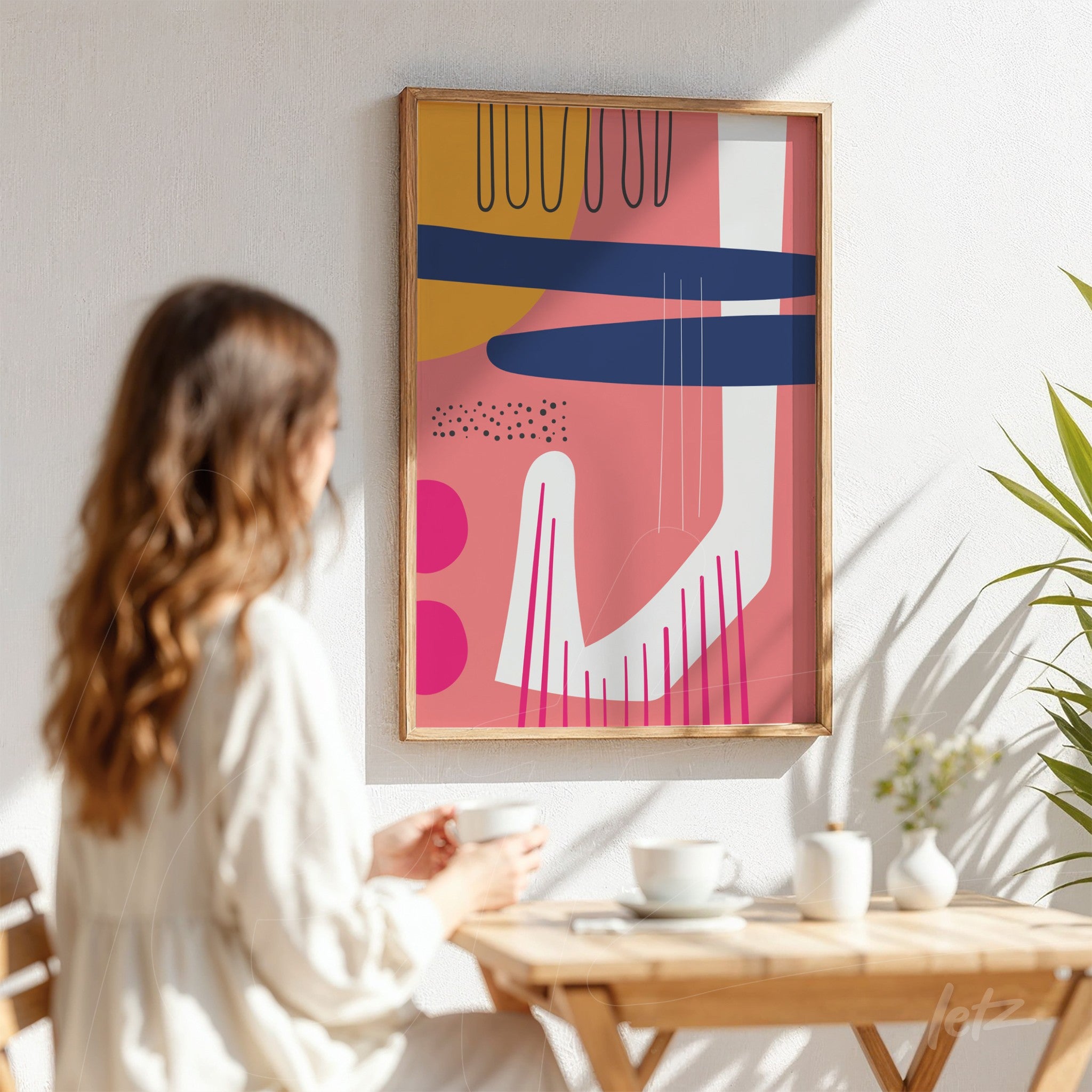 vista de quadro decorativo com arte boho em cores modernas, moldura de madeira clara, exibido em parede branca próximo a uma mulher sentada à mesa