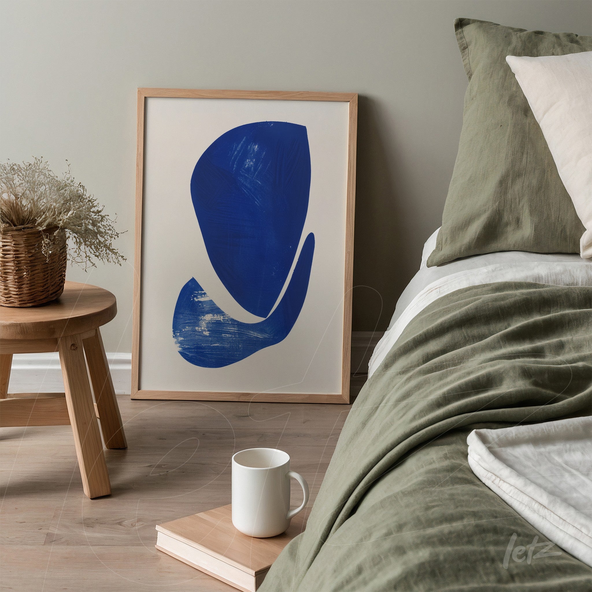 quadro emoldurado com arte abstrata em azul, apoiado ao lado de uma cama com roupa de cama em tons verde e beige
