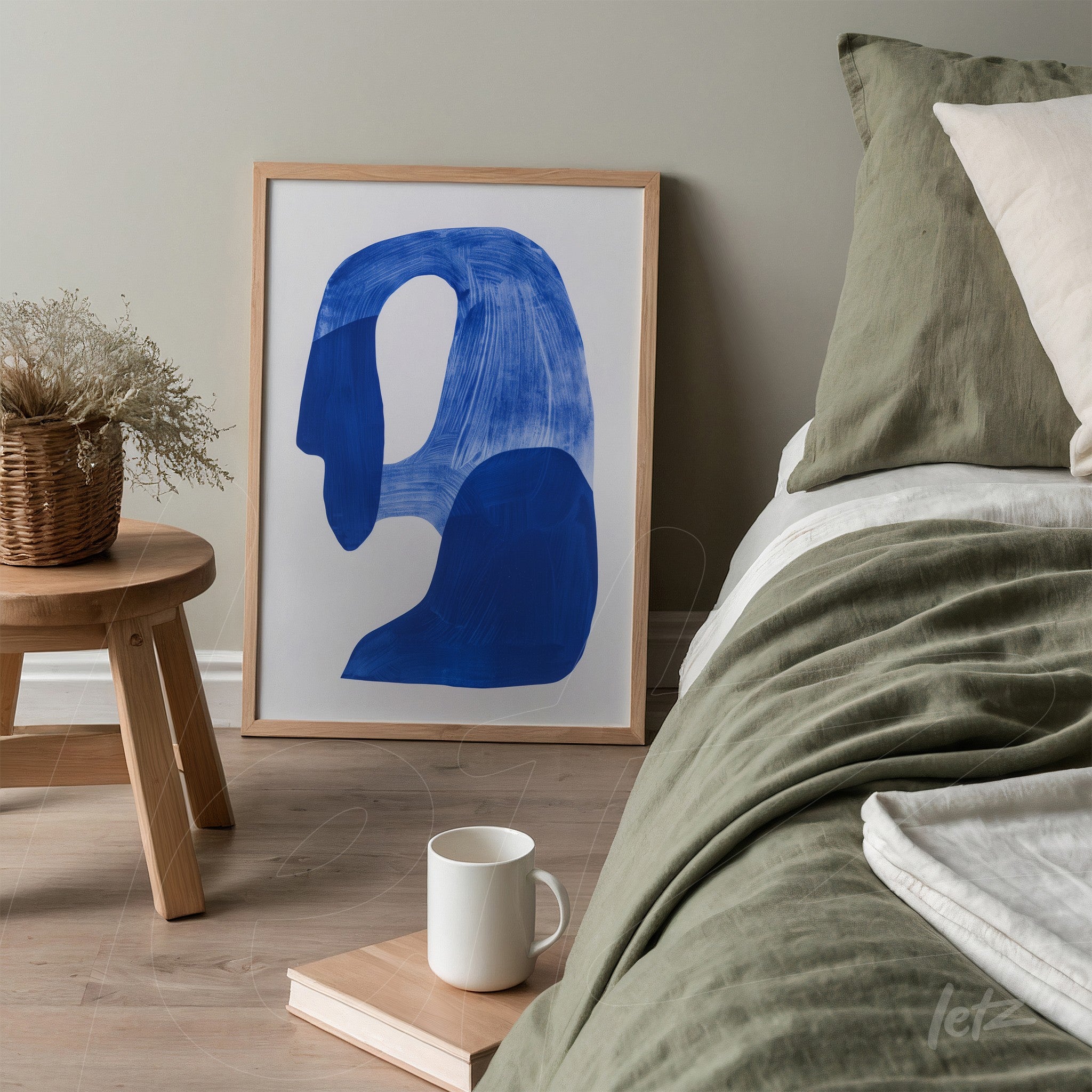 quadro com arte abstrata de formas em azul escuro em moldura clara, apoiado ao lado de cama com colcha verde