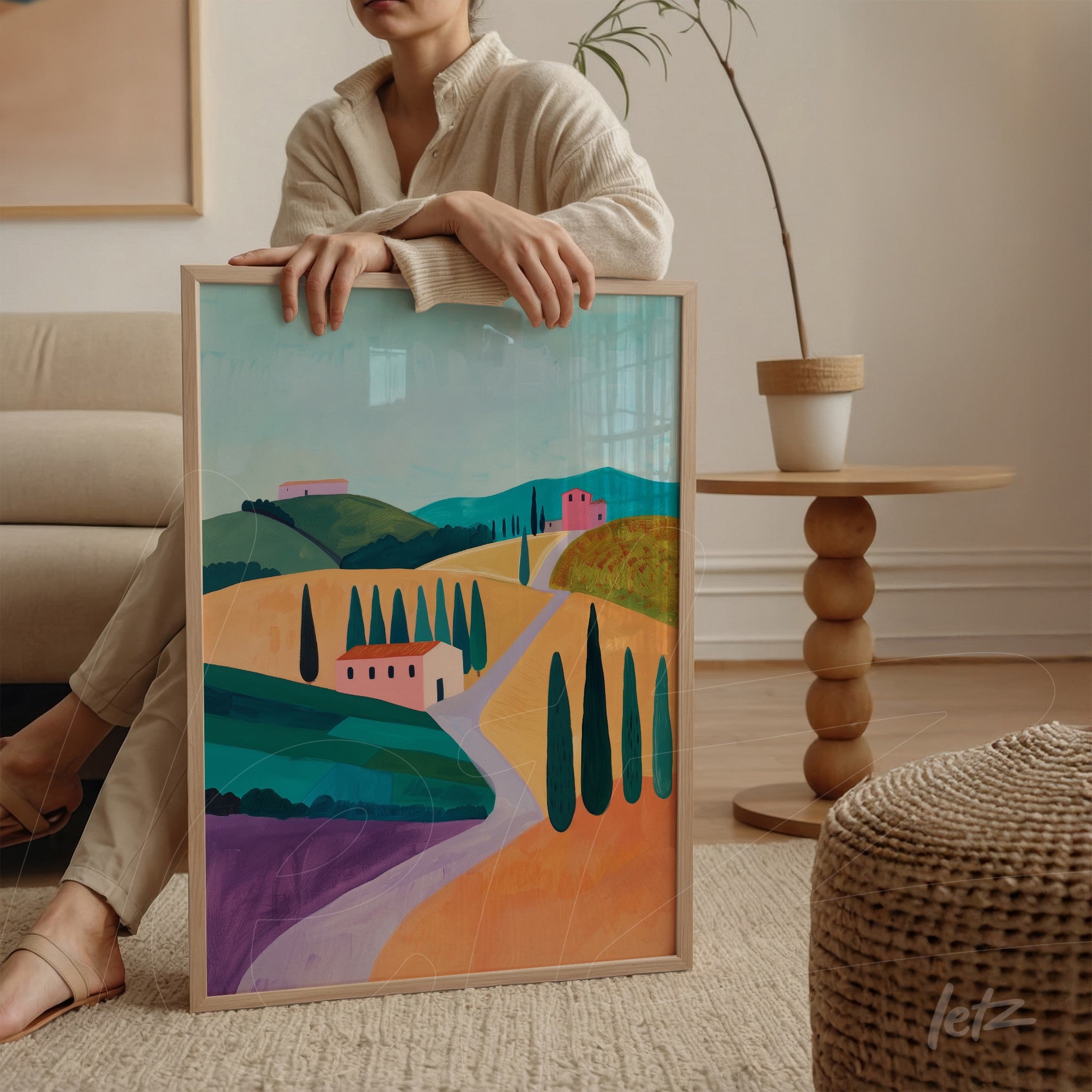 quadro em moldura clara com paisagem vibrante de colinas, vilarejos e ciprestes, segurado por mulher sentada em ambiente moderno