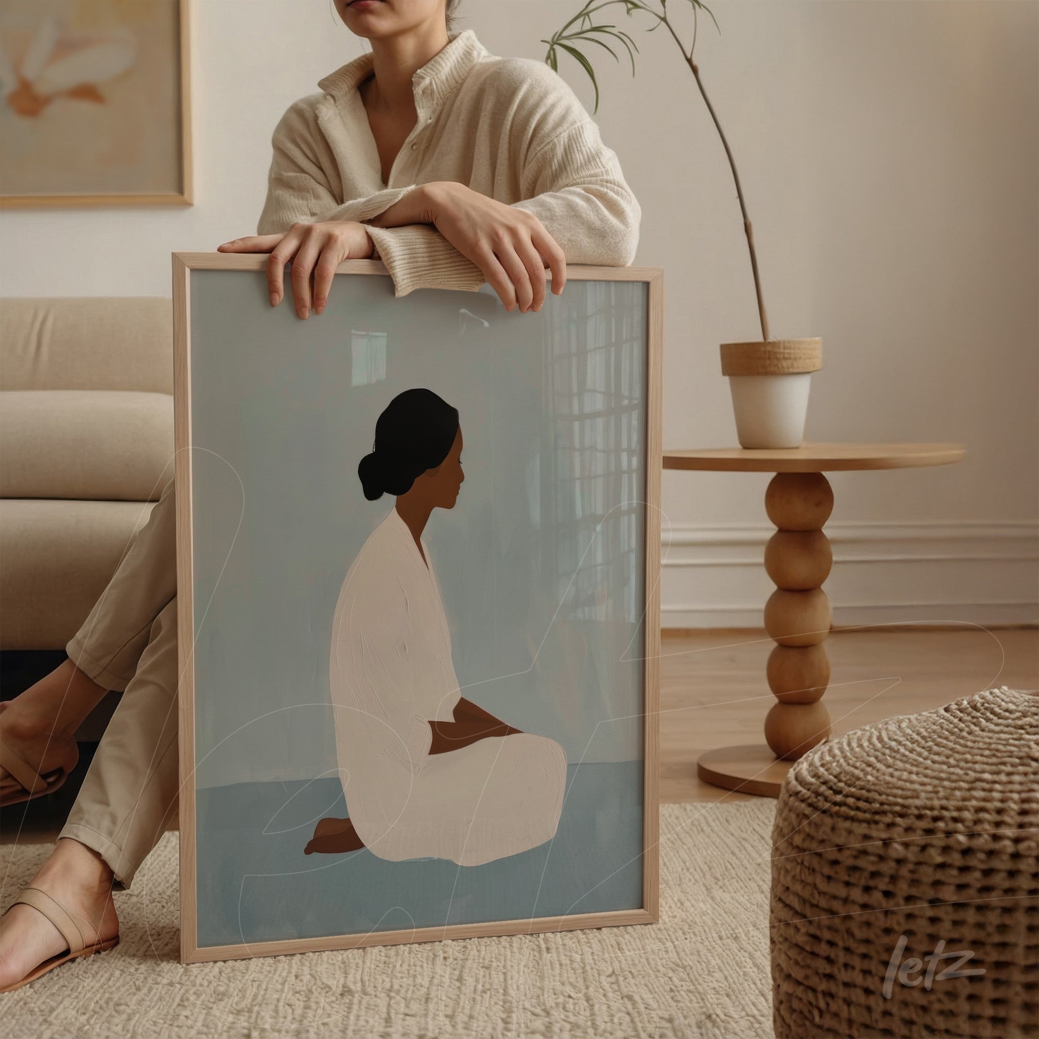 quadro com arte minimalista de figura feminina sentada em fundo azul, moldura de madeira clara, apoiado sobre mesa ao lado de uma mulher