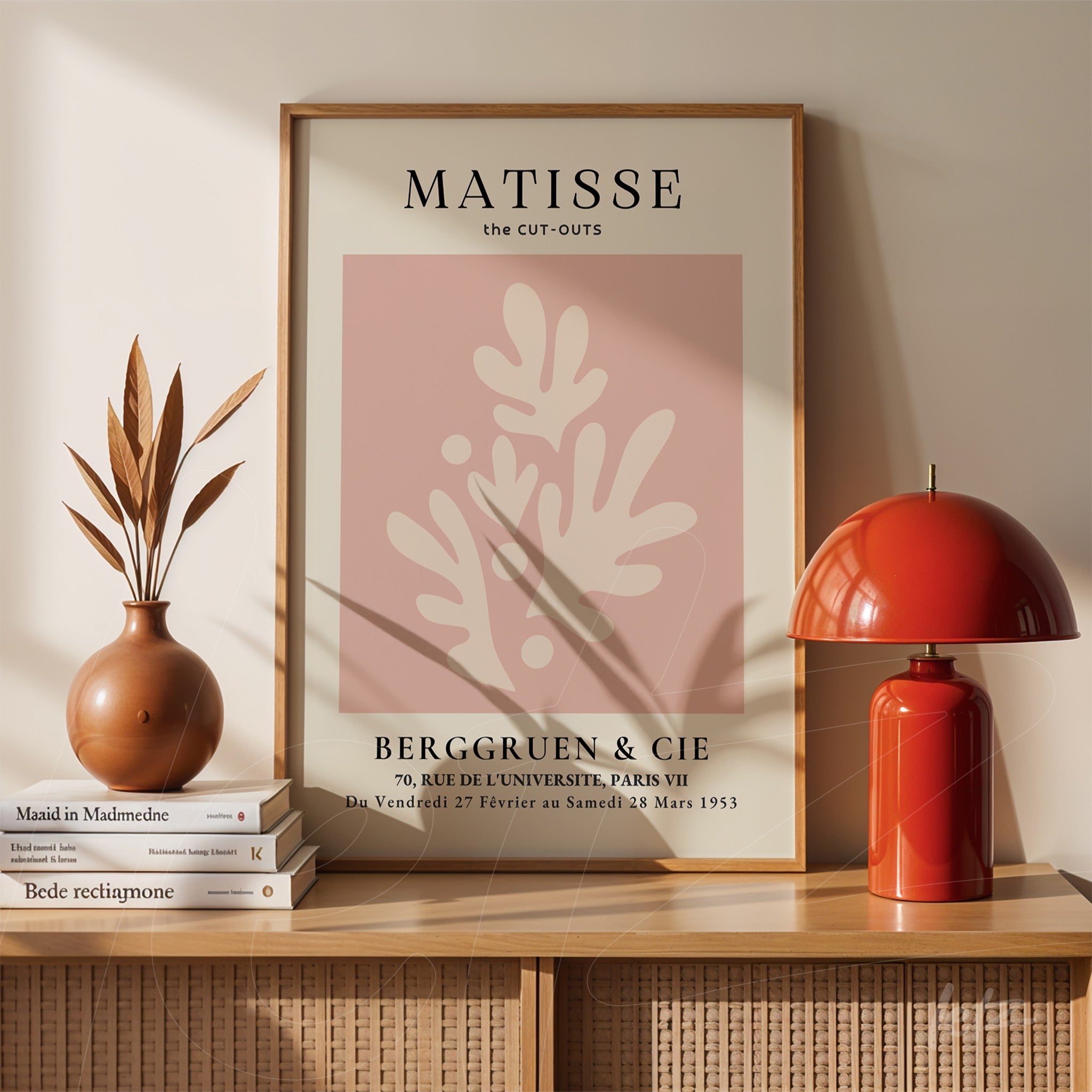 quadro com arte abstrata de folhas em fundo rosa, moldura de madeira clara e exposto sobre uma prateleira de madeira ao lado de um vaso e uma luminária