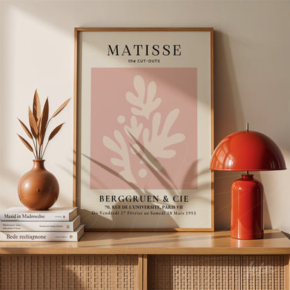 quadro com arte abstrata de folhas em fundo rosa, moldura de madeira clara e exposto sobre uma prateleira de madeira ao lado de um vaso e uma luminária