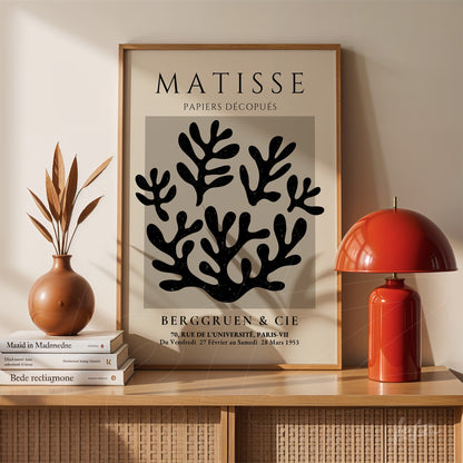 quadro decorativo com arte de folhas em preto sobre fundo cinza, moldura de madeira clara, apoiado em prateleira de madeira ao lado de vaso de cerâmica e luminária vermelha