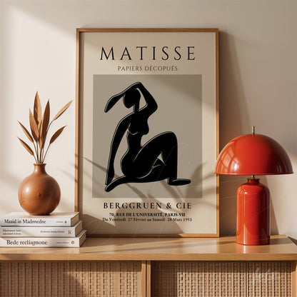 quadro com arte de figura humana estilizada em fundo cinza, moldura clara e suporte sobre mesa de madeira