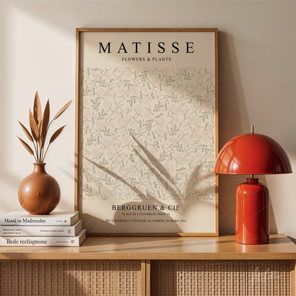 quadro com arte de plantas de matisse em moldura de madeira clara apoiado sobre uma mesa com vaso e luminária vermelha