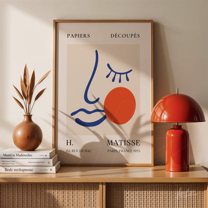 quadro decorativo em estilo minimalista com traços abstratos de rosto e círculo avermelhado, moldura de madeira clara exposto sobre mesa ao lado de abajur vermelho
