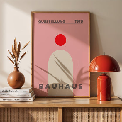 pôster de arte bauhaus com círculo vermelho e fundo rosa em moldura de madeira clara, exibido sobre móvel de madeira ao lado de uma lâmpada vermelha e vaso com folhas secas