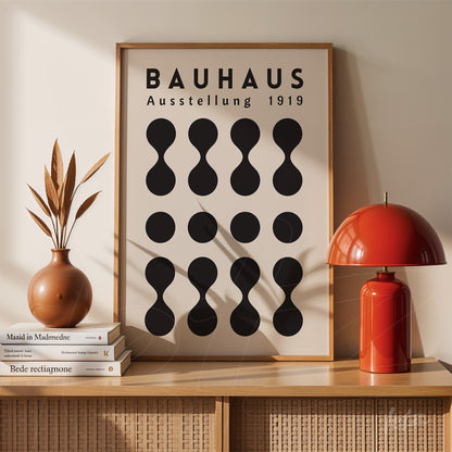 pôster impresso com design moderno de formas geométricas pretas sob fundo bege e texto 'Bauhaus Ausstellung 1919', emoldurado e apoiado em aparador de madeira claro ao lado de lâmpada vermelha