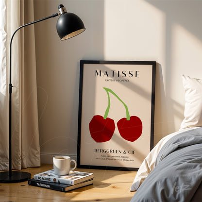 quadro em moldura preta com arte moderna de cerejas em estilo minimalista, exibido ao lado de um abajur sobre mesa de madeira e perto de uma cama com roupa de cama clara