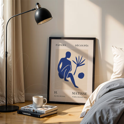 quadro emoldurado com arte estilizada de figuras humanas em azul sobre fundo claro, exibido ao lado de uma luminária de pé e pilha de livros em uma mesa de madeira