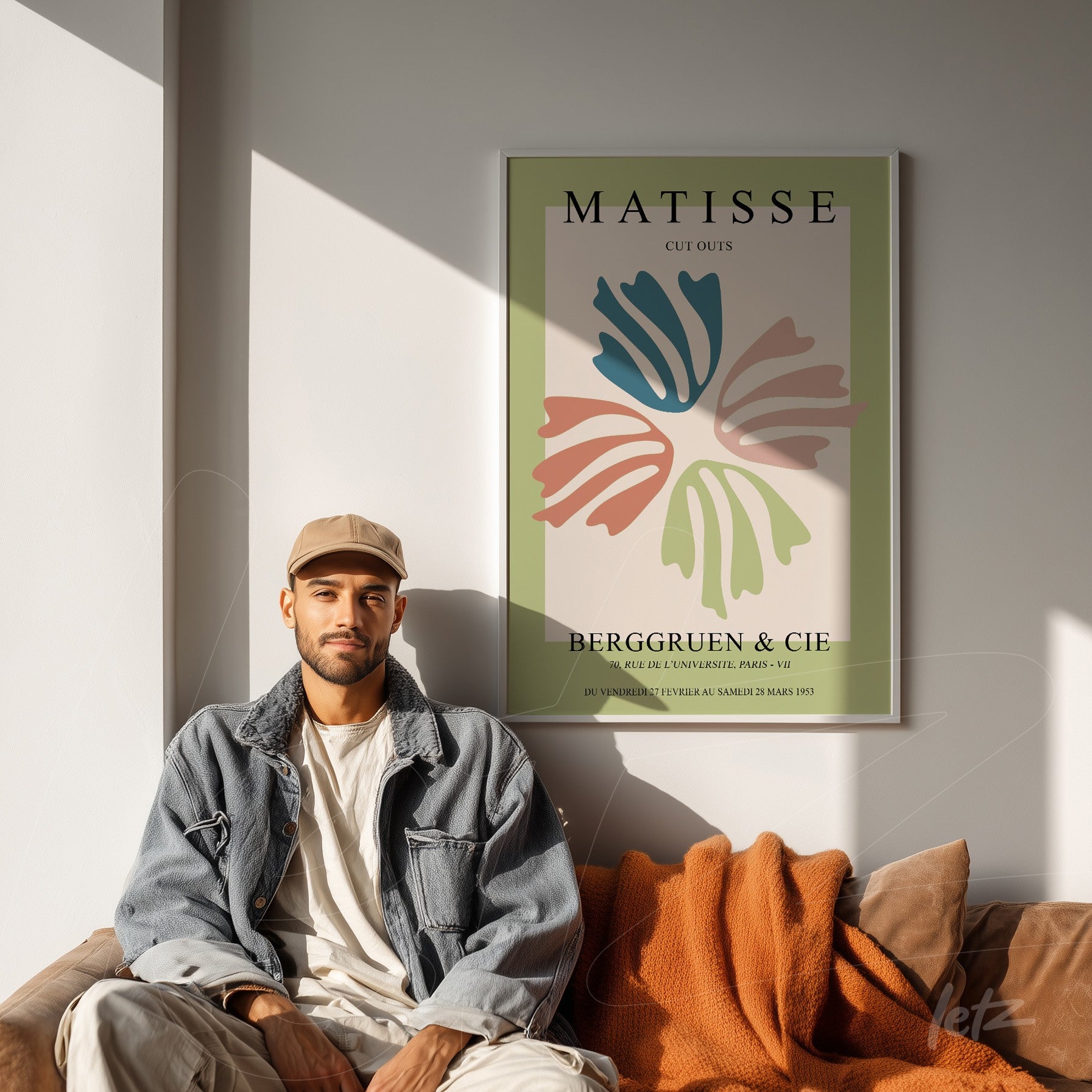 quadro de arte moderna com ilustrações em cores suaves de folhas em moldura fina sobre parede clara, exibido ao lado de um jovem homem sentado em um sofá com cobertor laranja