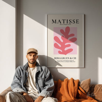 quadro com arte moderna de fundo rosa e folhas em tons de vermelho, moldura branca, exposto em parede iluminada com sofá marrom e manta laranja