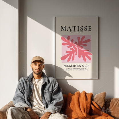 quadro em moldura branca com arte moderna de Matisse, exibindo folhas em tons de rosa, apresentado acima de um sofá com cobertor laranja e uma pessoa sentada ao lado