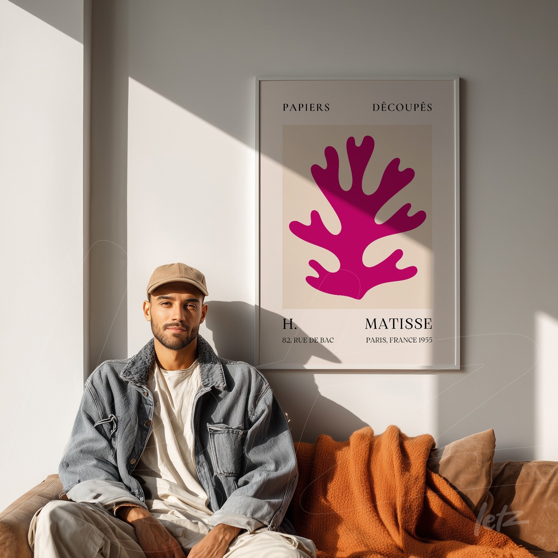 quadro emoldurado com arte gráfica de um coral rosa sobre fundo claro, exibido em sala iluminada por luz natural, acompanhado por um jovem sentado em sofá com almofadas laranjas