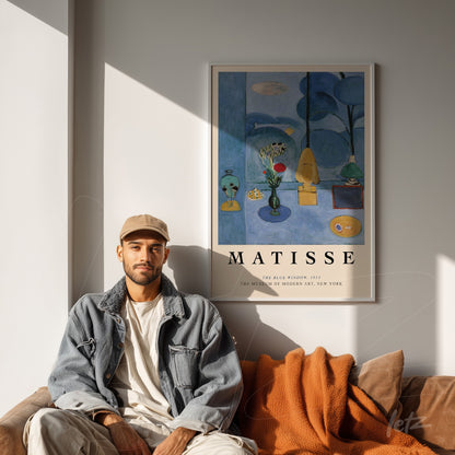 quadro de arte moderna na parede com moldura clara e presença de elementos como vasos verdes, flores e uma tela colorida de Matisse, exibido em ambiente aconchegante com sofá marrom e cobertor laranja