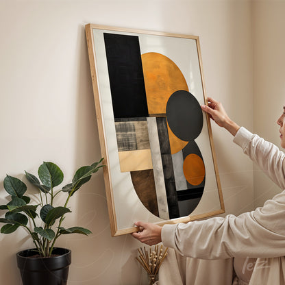 quadro com arte abstrata em cores laranja e preto, moldura de madeira clara, sendo segurado por uma pessoa em ambiente com planta