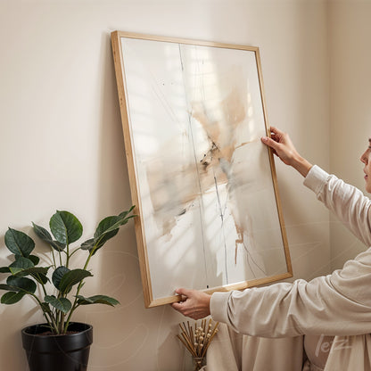 quadro abstrato com tons neutros em moldura de madeira clara, sendo segurado em um ambiente com planta