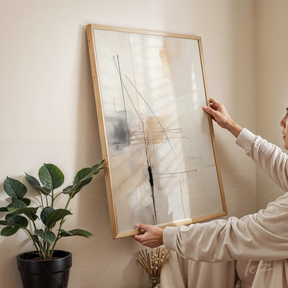 quadro abstrato em moldura de madeira clara com linhas e formas sutis em tons neutros, segurado por pessoa ao lado de planta verde