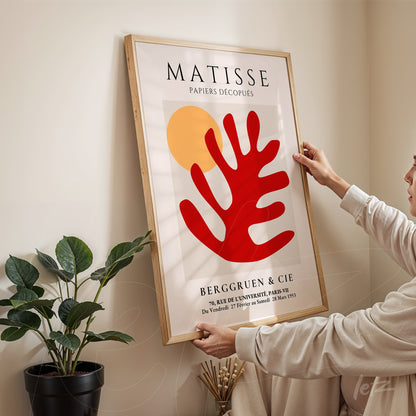 quadro decorativo com arte abstrata em tons vermelho e amarelo, com moldura de madeira clara, exibido sobre parede neutra