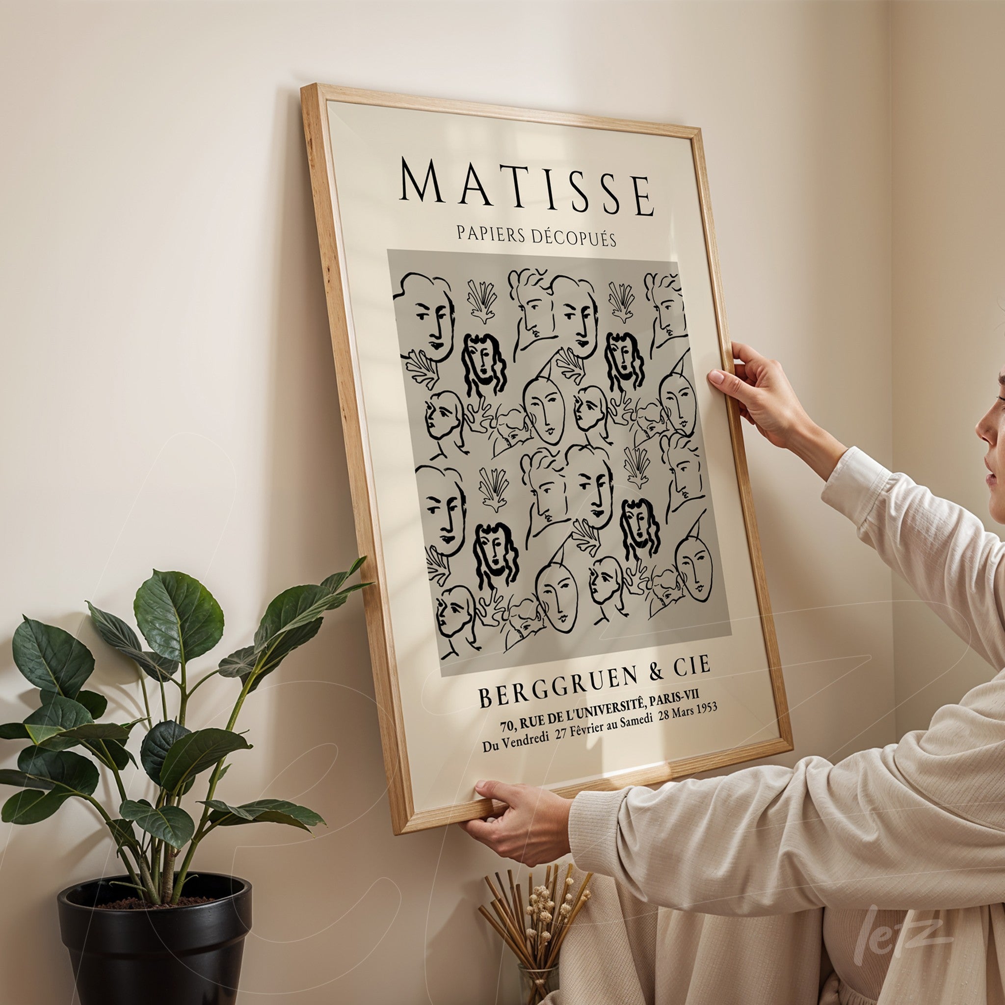 quadro decorativo com ilustração em preto e branco de rostos em estilo minimalista, moldura de madeira clara, pendurado em parede com planta ao lado