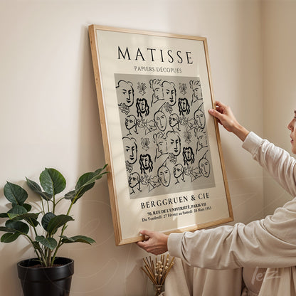 quadro decorativo com ilustração em preto e branco de rostos em estilo minimalista, moldura de madeira clara, pendurado em parede com planta ao lado