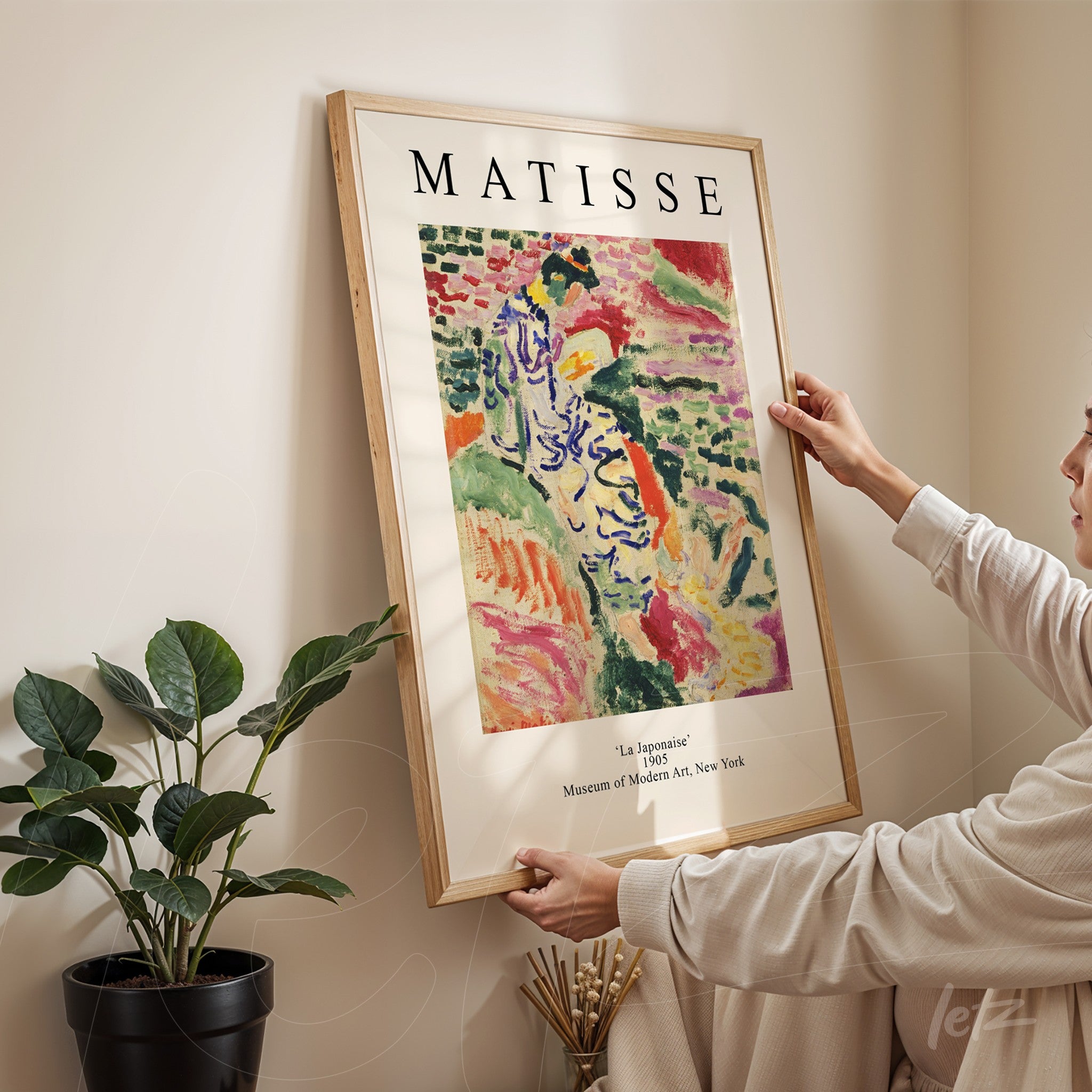 quadro com arte de Matisse, intitulado 'La Jalousie', em moldura de madeira clara, sendo segurado por uma pessoa em ambiente interno com planta ao fundo