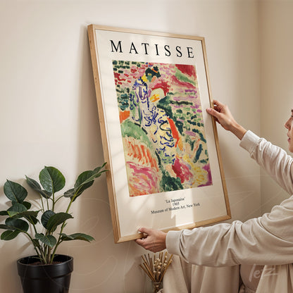 quadro com arte de Matisse, intitulado 'La Jalousie', em moldura de madeira clara, sendo segurado por uma pessoa em ambiente interno com planta ao fundo