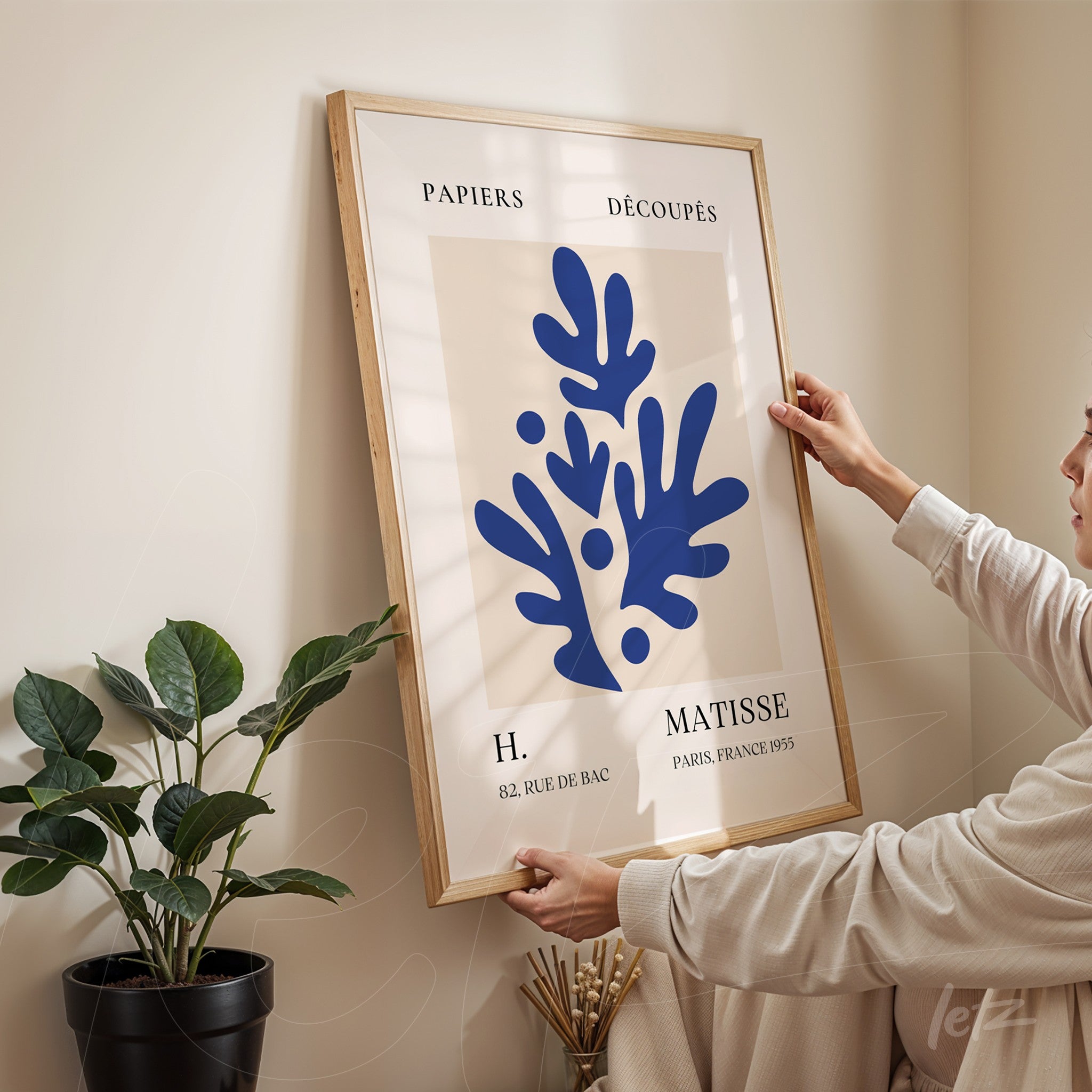 quadro com arte em estilo moderno apresentando formas abstratas em azul, emoldurado em madeira clara, sendo segurado por uma pessoa diante de parede clara