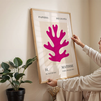 quadro com arte contemporânea de forma coral em tons de rosa e moldura de madeira clara exibido em ambiente interno com planta ao fundo