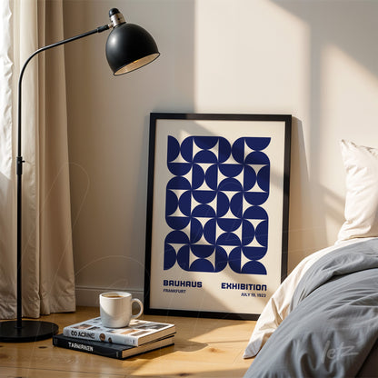 quadro com arte geométrica azul sobre fundo branco apoiado em uma parede clara ao lado de uma cama, com abajur preto de chão ao lado