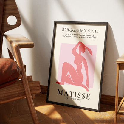 quadro com arte abstrata em tons de rosa e vermelho, mulher estilizada em fundo claro, moldura preta fina, apoiado em cadeira de madeira