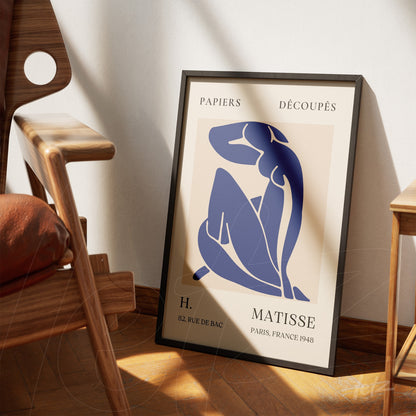 quadro com arte em estilo minimalista de figura feminina em azul, apoiado em parede clara ao lado de uma cadeira moderna