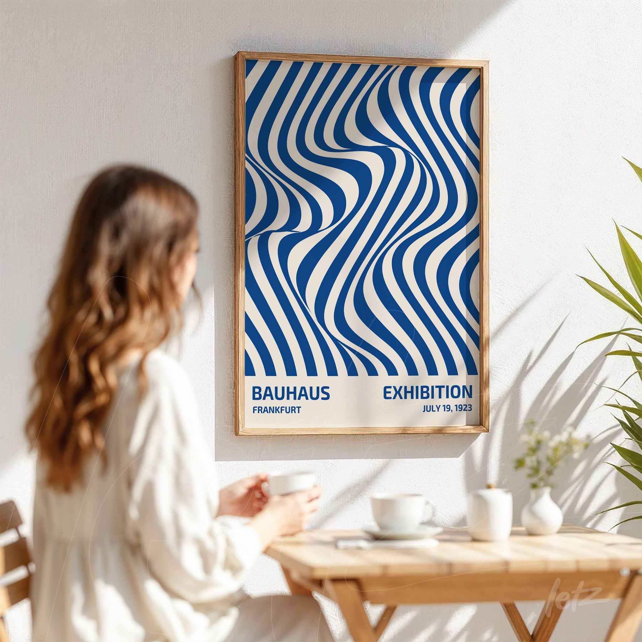pôster com design de linhas abstratas sinuosas em azul e branco em moldura de madeira clara apoiado em parede branca com uma mulher ao lado