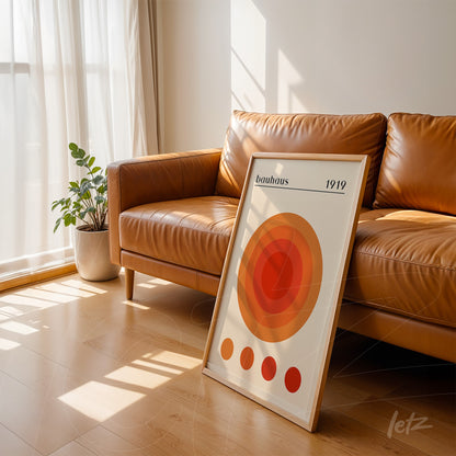 quadro com arte geométrica estilo bauhaus com círculos concêntricos em tons de laranja, apoiado em um sofá de couro marrom em ambiente iluminado