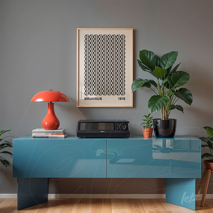 pôster com padrão geométrico em moldura de madeira clara sobre parede cinza, ao lado de luminária moderna vermelha e plantas verdes