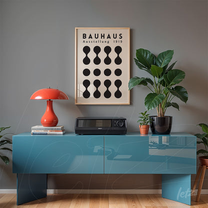 pôster com design geométrico preto e branco em moldura clara exibido sobre parede cinza, acompanhado de plantas e luminária vermelha