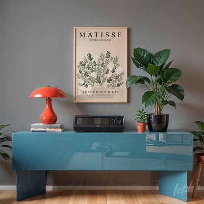 quadro decorativo com arte de Matisse em moldura clara exibido em parede cinza, com mesa azul e planta ao fundo