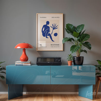 quadro com arte de Henri Matisse em moldura dourada, exposto sobre um móvel azul com lampe de mesa laranja e planta em vaso preto ao lado