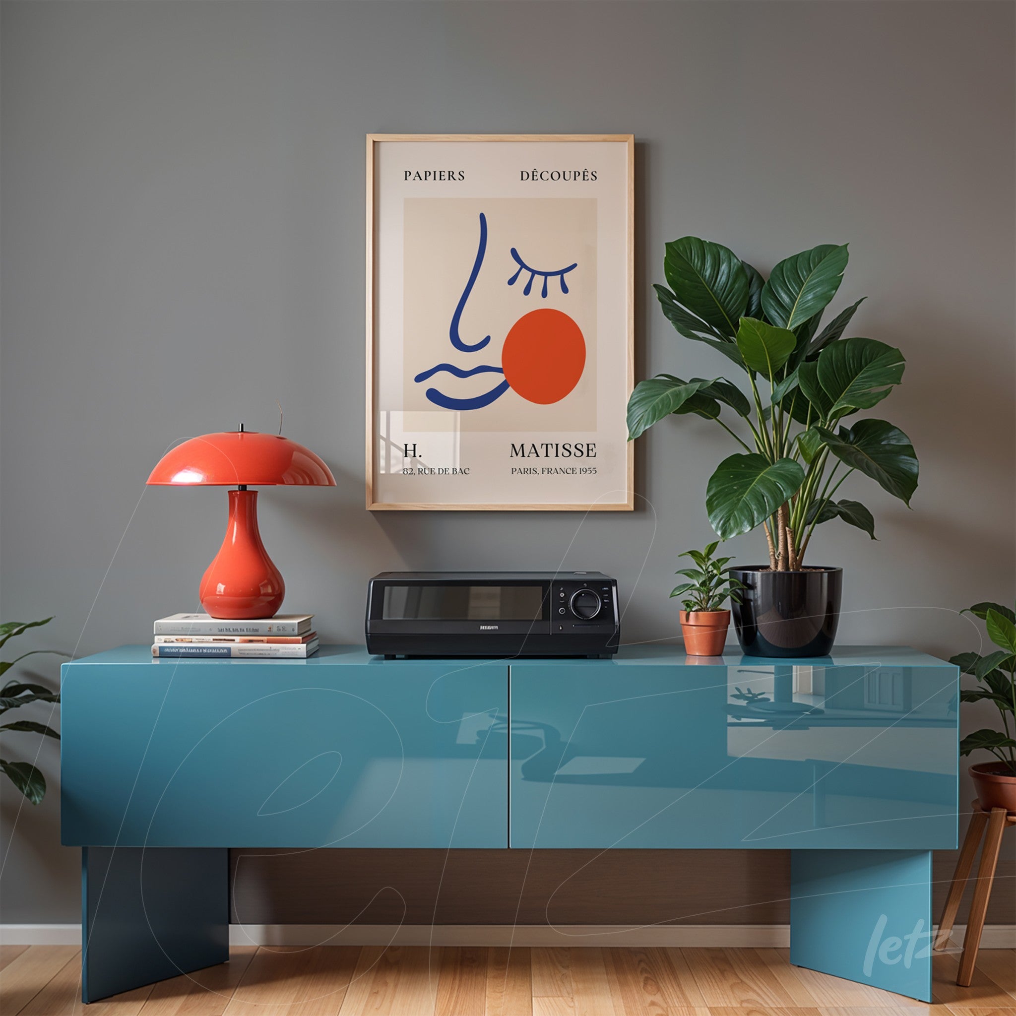 quadro decorativo em moldura dourada com ilustração minimalista de rosto e detalhes em vermelho, exposto sobre uma mesa azul com um vaso de planta ao lado