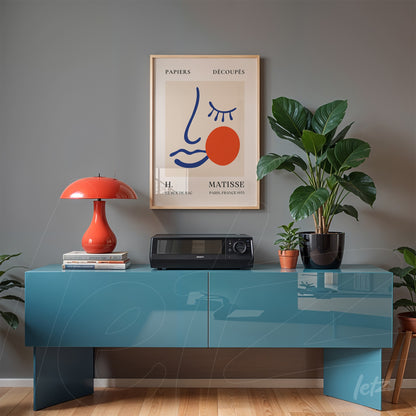 quadro decorativo em moldura dourada com ilustração minimalista de rosto e detalhes em vermelho, exposto sobre uma mesa azul com um vaso de planta ao lado