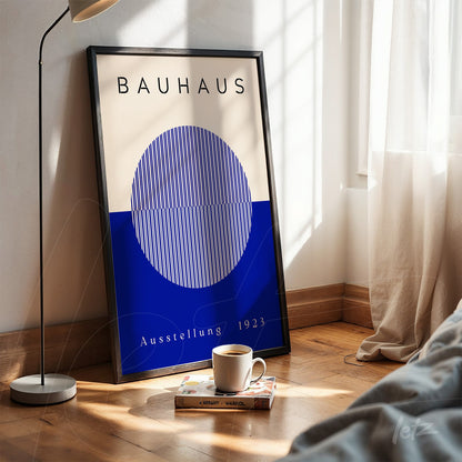 pôster impresso de design bauhaus com esfera azul e linhas brancas em moldura preta, apoiado no chão de madeira ao lado de xícara de café