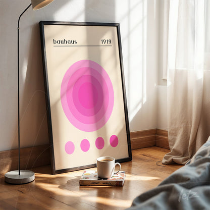 pôster impresso com círculos concêntricos em rosa sob fundo bege, apoiado em piso de madeira com moldura preta, luz natural à esquerda