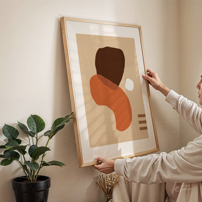 arte abstrata em tons terrosos com formas orgânicas exibida em moldura de madeira clara, segurada por uma pessoa e apoiada em parede creme ao lado de uma planta verde em vaso preto